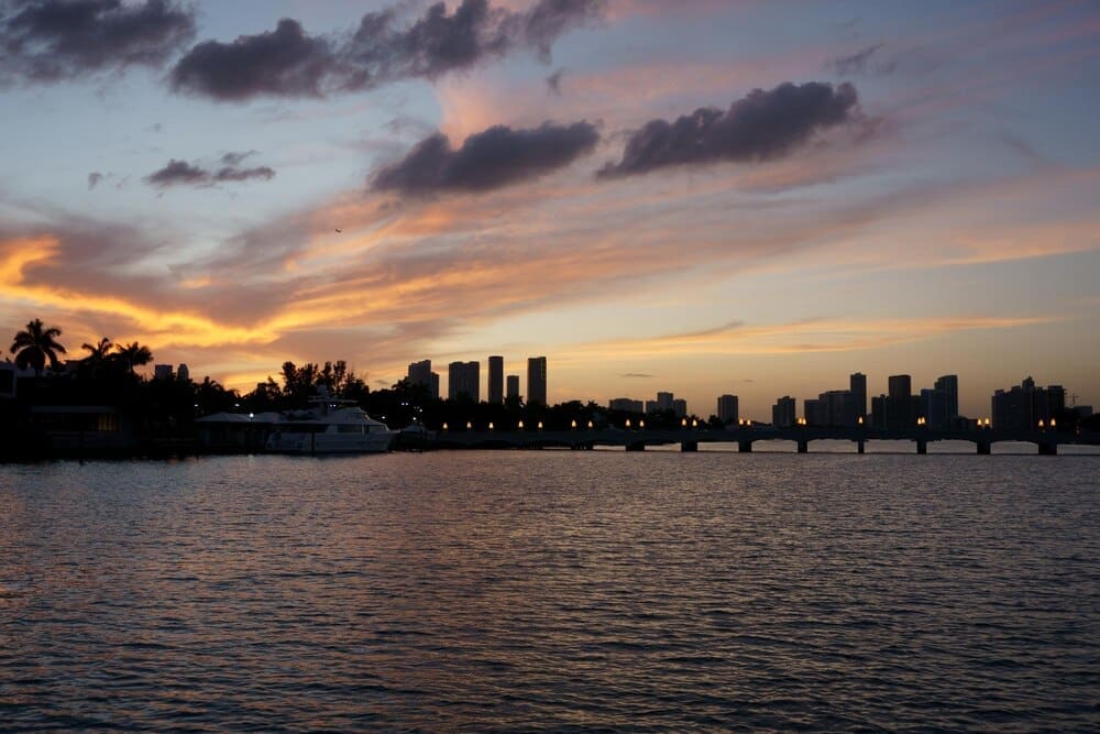 Sunset illuminates Biscayne Bay flowing past buildings, silhouetted against the colorful sky.