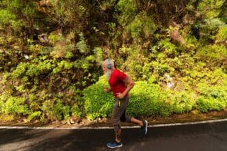 A senior man with a white beard runs outdoors in a natural setting.