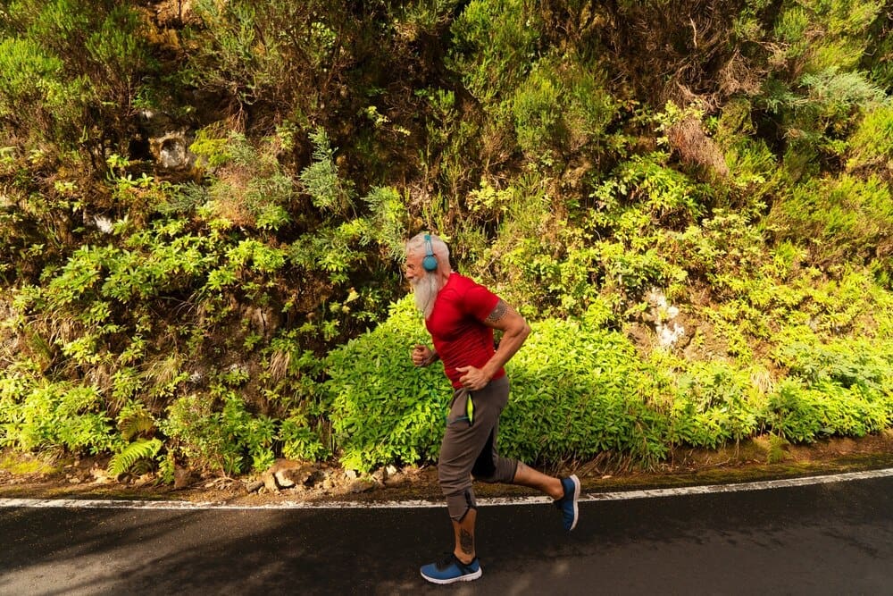 A senior man with a white beard runs outdoors in a natural setting.
