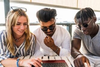 A diverse group of friends laughs while looking at a laptop screen outdoors, suggesting a summer vacation.