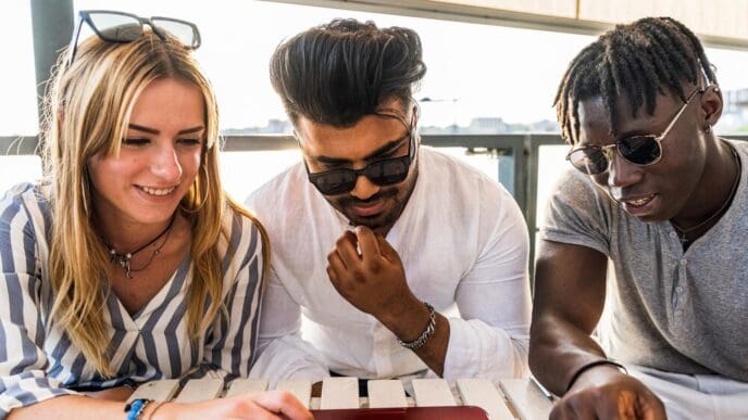 A diverse group of friends laughs while looking at a laptop screen outdoors, suggesting a summer vacation.