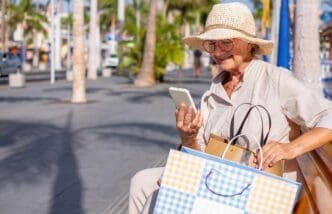 Smiling senior woman in a straw hat sits on a city bench, reading a message on her smartphone.