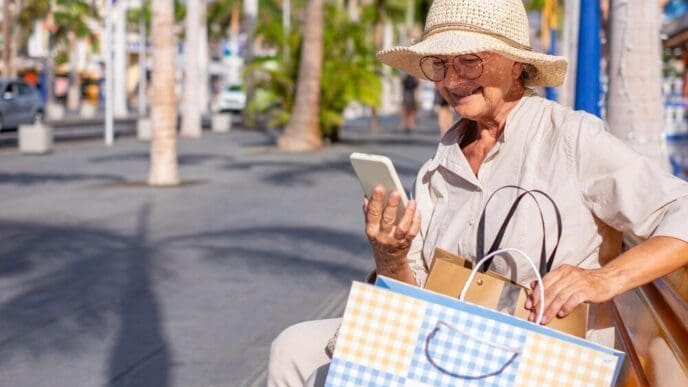 Smiling senior woman in a straw hat sits on a city bench, reading a message on her smartphone.