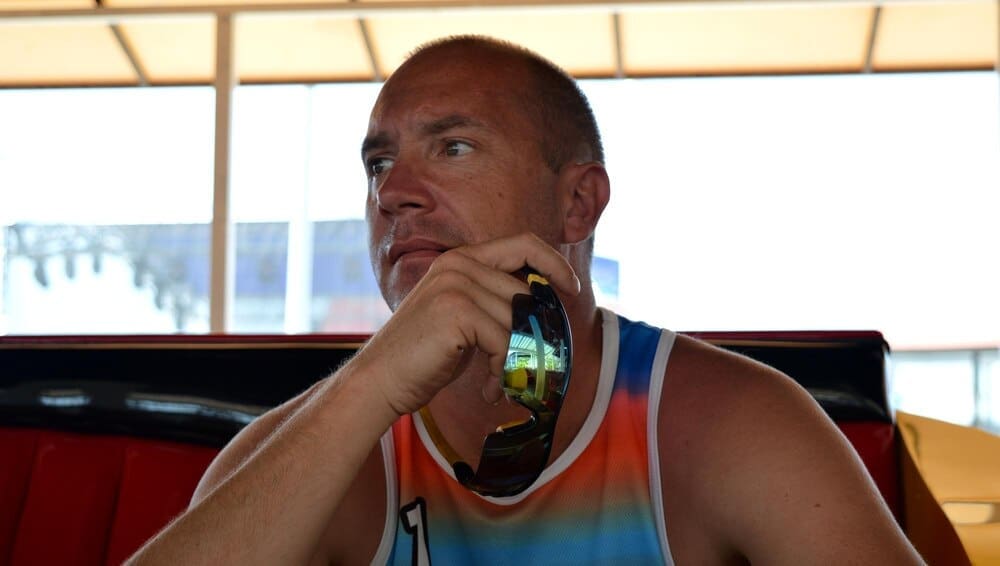A man with a thoughtful expression sits on a sofa, holding sunglasses.