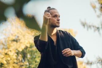 A young man stands against a backdrop of plants, gazing thoughtfully away from the camera.
