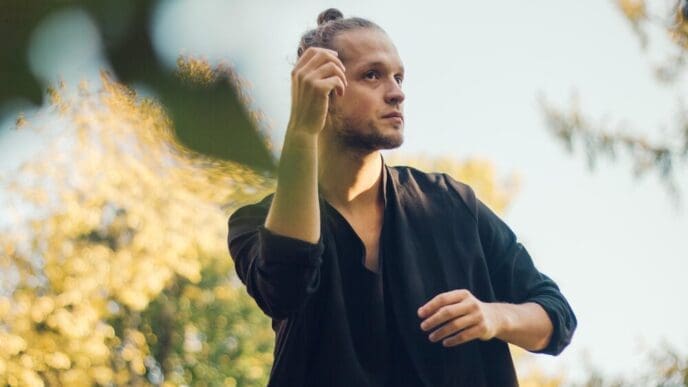 A young man stands against a backdrop of plants, gazing thoughtfully away from the camera.