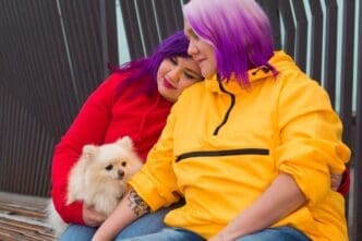 A woman with two-toned hair sits on an outdoor bench, holding a small, fluffy dog.