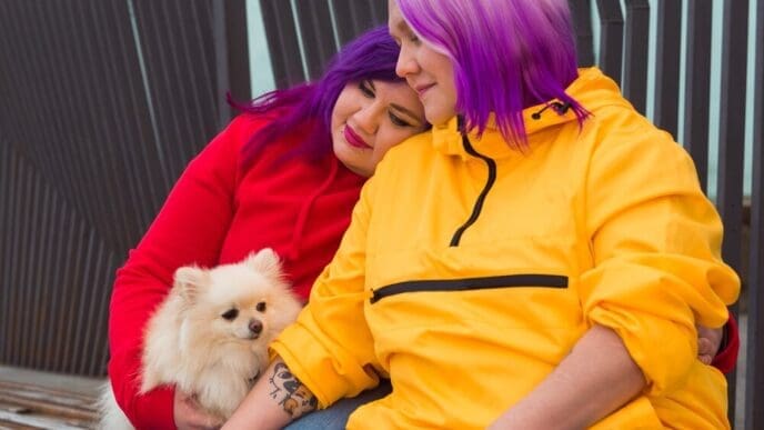 A woman with two-toned hair sits on an outdoor bench, holding a small, fluffy dog.