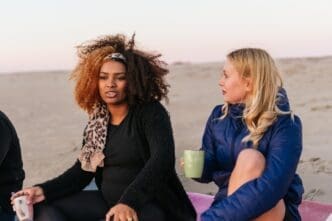 Two women of diverse ethnicities enjoy tea and conversation while seated on a beach at sunset.