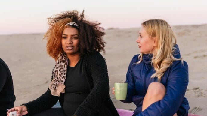 Two women of diverse ethnicities enjoy tea and conversation while seated on a beach at sunset.
