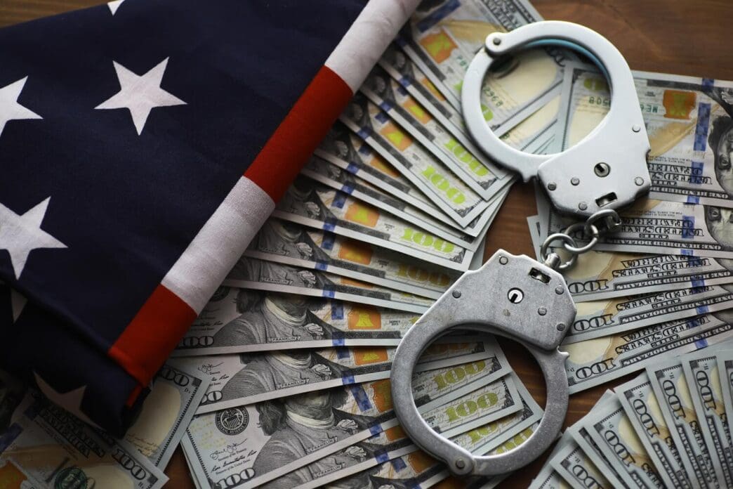 A pair of handcuffs rests on a U.S. flag, with money scattered around, illustrating concepts of law enforcement and financial crime.