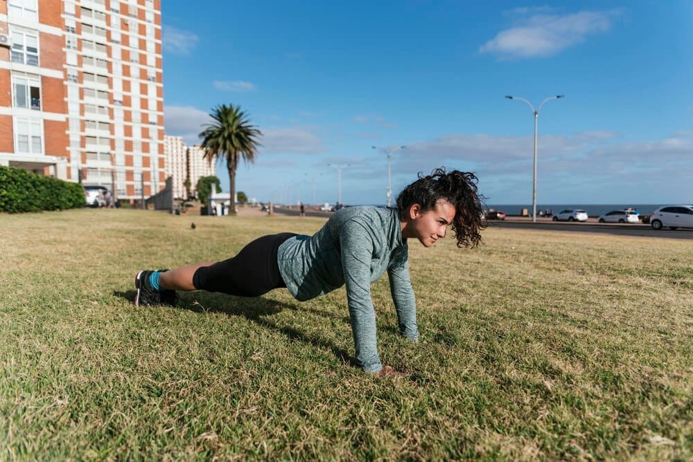 A woman in workout clothes performs push-ups on a grassy surface.