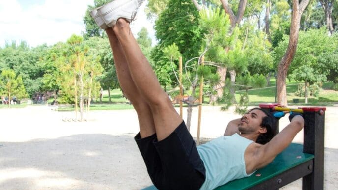 A woman lies on a bench in a park and performs a leg lift.