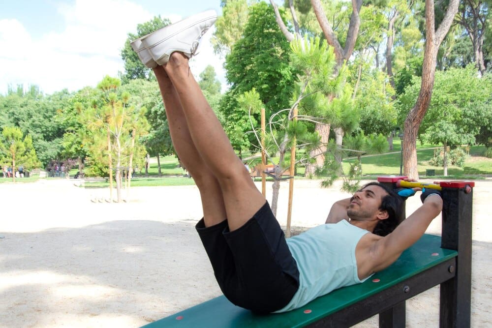 A woman lies on a bench in a park and performs a leg lift.