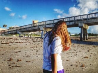 Woman stands on the sand in front of a pier on a sunny beach.
