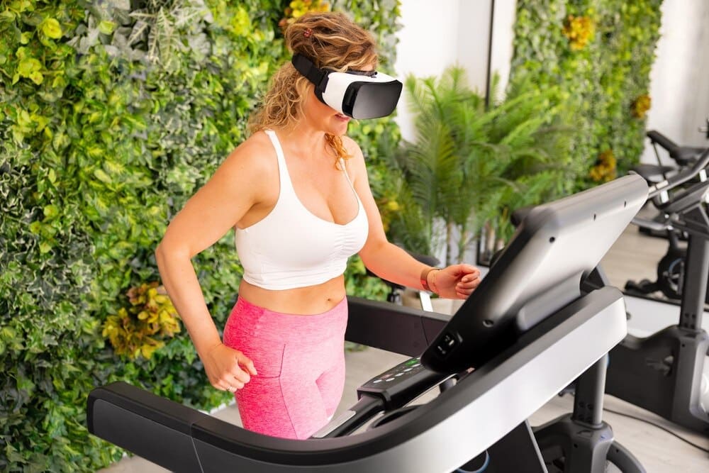 A woman wearing a VR headset runs on a treadmill in a gym.