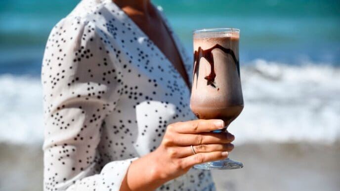 A woman in a white dress holds a coffee latte while standing on a beach in Greece.