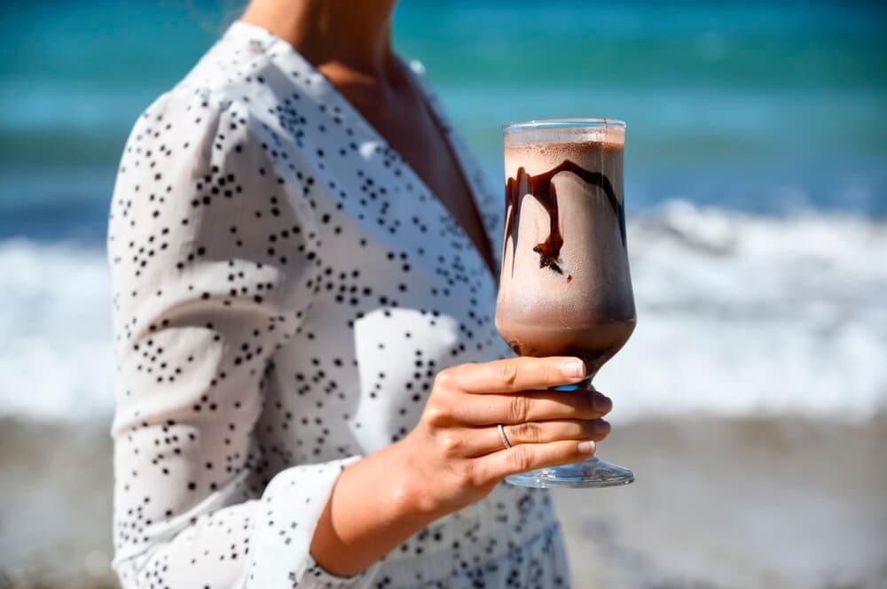 A woman in a white dress holds a coffee latte while standing on a beach in Greece.