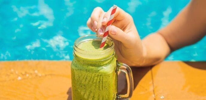 Woman holding a green spinach and banana smoothie with a pool in the background.