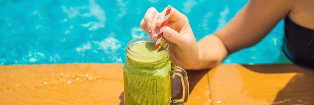 Woman holding a green spinach and banana smoothie with a pool in the background.
