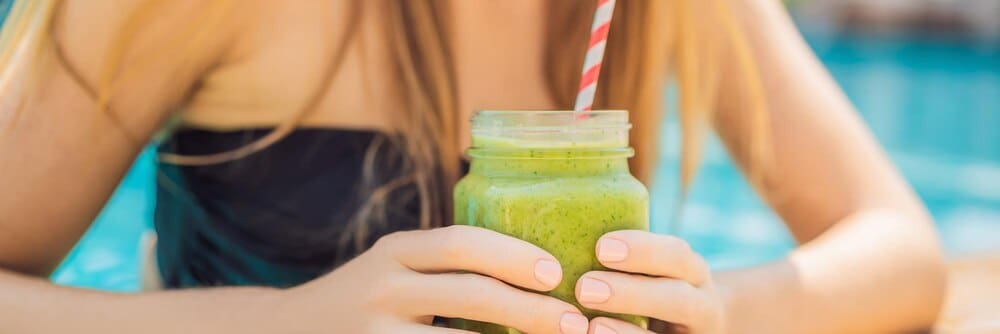 A woman holds a green smoothie made with spinach and banana near a swimming pool, representing healthy eating.