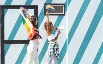 A woman speaks into a megaphone, with a rainbow LGBTQ+ pride flag visible in the background.
