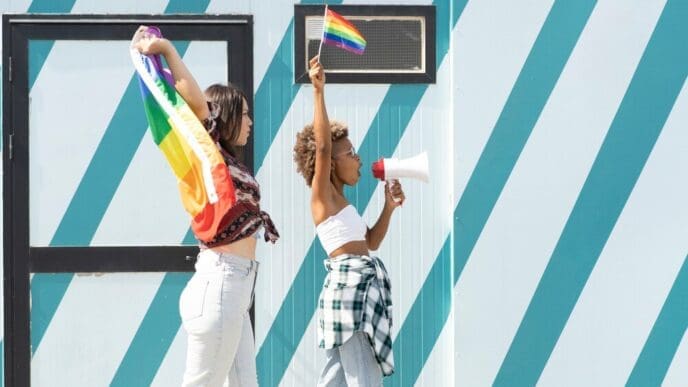 A woman speaks into a megaphone, with a rainbow LGBTQ+ pride flag visible in the background.