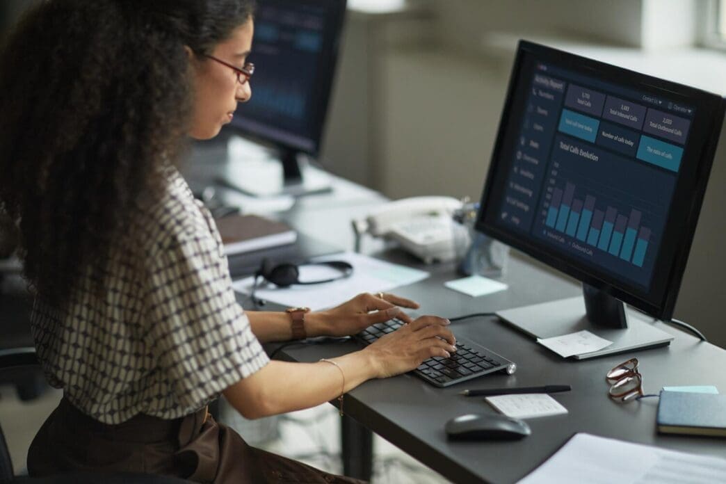 A young adult woman with dark hair analyzes financial data displayed on a computer monitor.