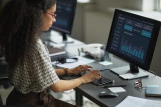 A young adult woman with dark hair analyzes financial data displayed on a computer monitor.