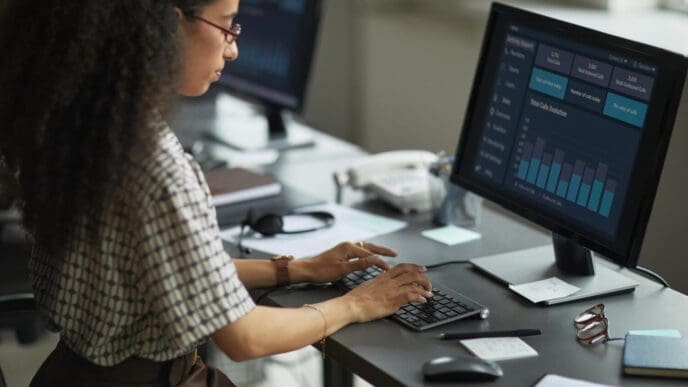A young adult woman with dark hair analyzes financial data displayed on a computer monitor.