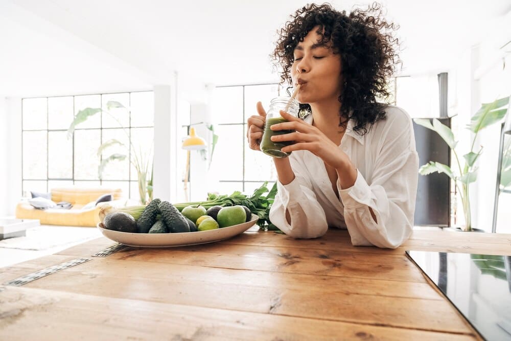 Young Black woman in a loft apartment drinks green juice from a bamboo straw, promoting a healthy lifestyle.