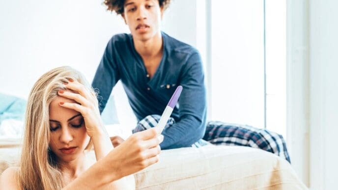 A young, interracial couple lies in bed, appearing unhappy about a pregnancy.