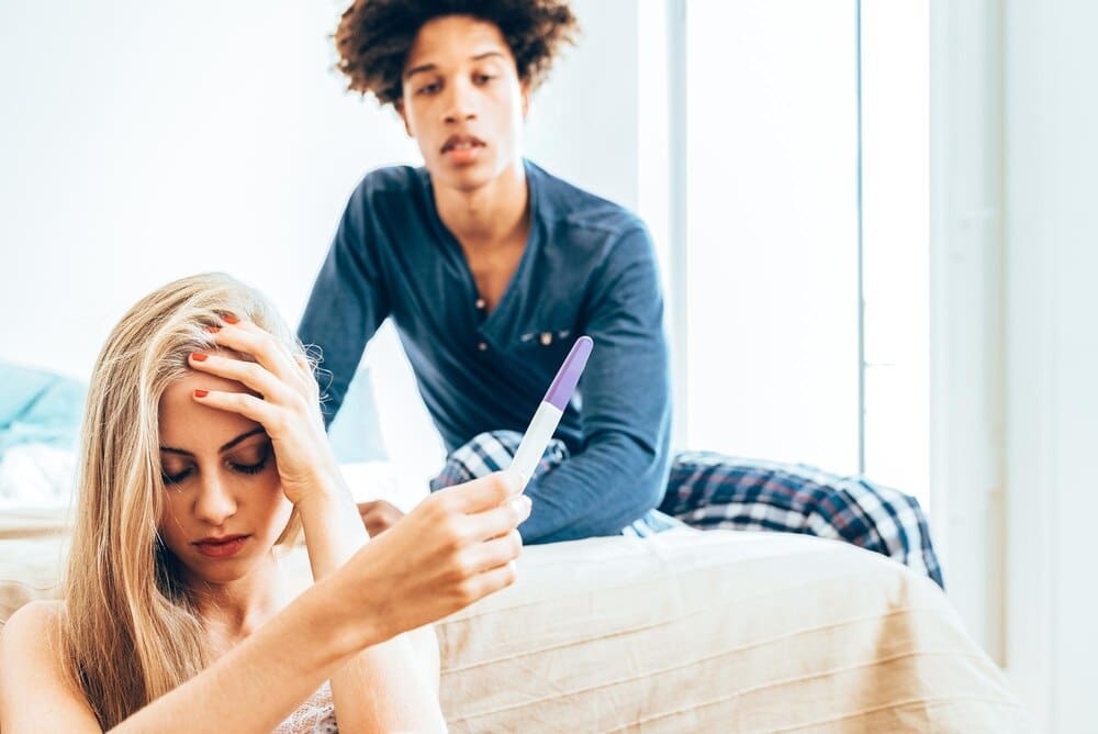 A young, interracial couple lies in bed, appearing unhappy about a pregnancy.