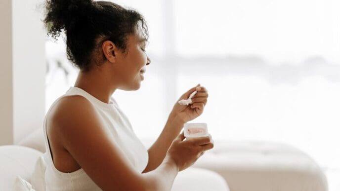 A young Black woman smiles while eating yogurt in her sunny kitchen.