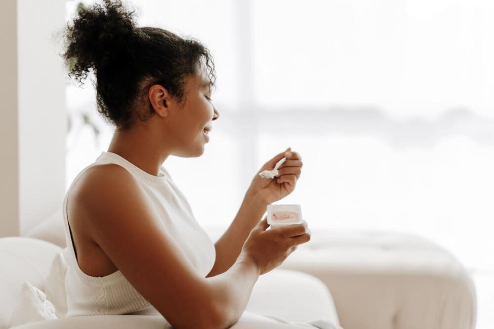 A young Black woman smiles while eating yogurt in her sunny kitchen.