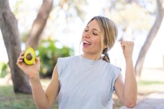 A young blonde woman celebrates outdoors, holding an avocado.