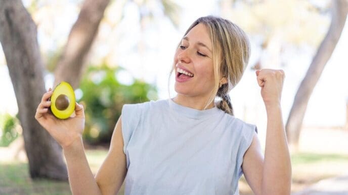 A young blonde woman celebrates outdoors, holding an avocado.