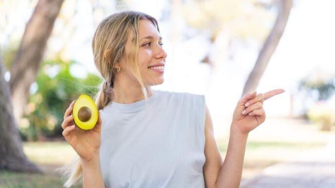 A young blonde woman outdoors holds an avocado and points to the side, likely presenting a product.