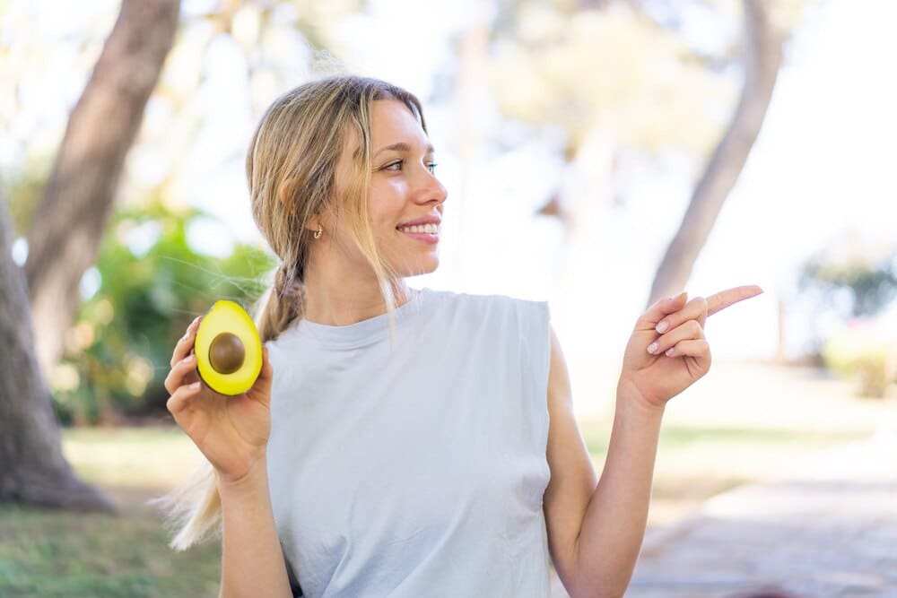 A young blonde woman outdoors holds an avocado and points to the side, likely presenting a product.