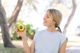 A young blonde woman smiles while holding an avocado outdoors.