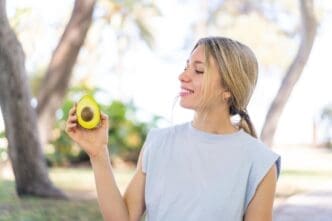 A young blonde woman smiles while holding an avocado outdoors.