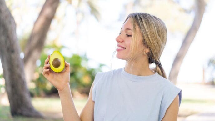 A young blonde woman smiles while holding an avocado outdoors.