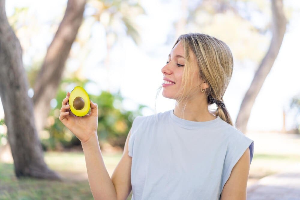 A young blonde woman smiles while holding an avocado outdoors.