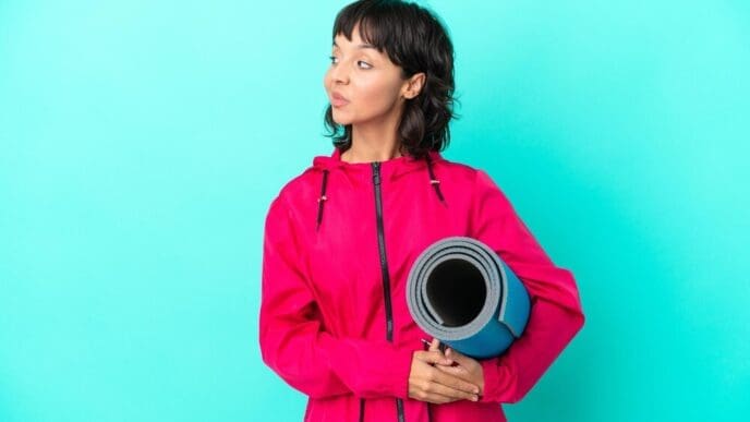 A young girl with a yoga mat stands on a blue background, looking to the side.
