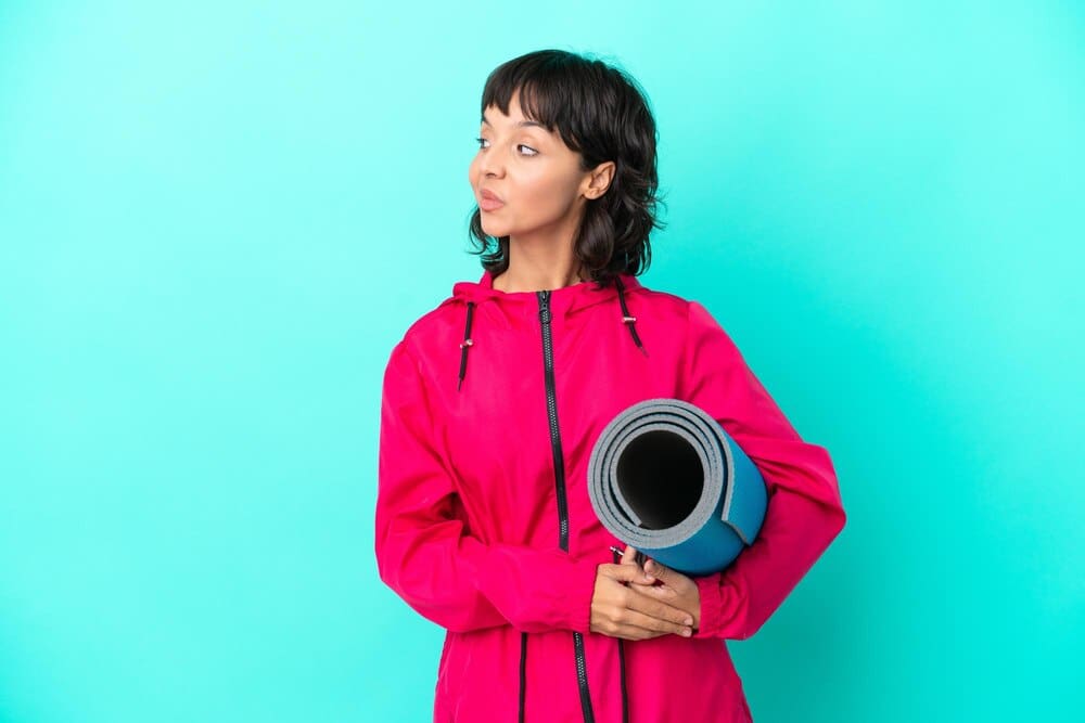 A young girl with a yoga mat stands on a blue background, looking to the side.