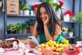 Young Hispanic woman, a florist, smiles confidently while using a smartphone in her flower shop.