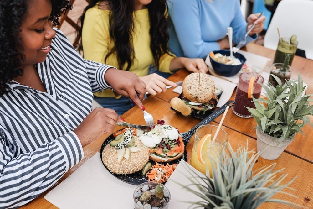 A young Black girl's right hand rests on a table, likely as she enjoys breakfast with friends on an outdoor restaurant terrace.