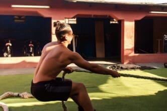 Young, shirtless man with defined muscles stretches a rope in an outdoor gym.