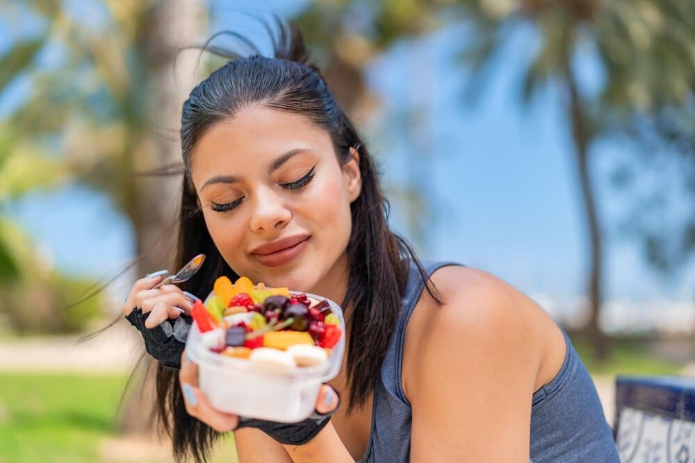 A young woman smiles while holding a bowl of fruit outside.