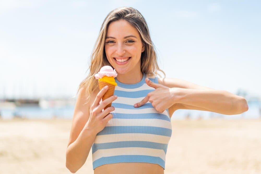 A young woman with a surprised expression holds a cornet ice cream cone outdoors.